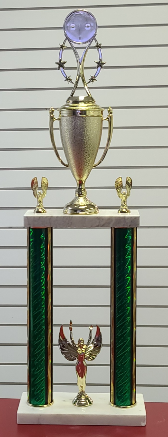 Tall two-column trophy with green holographic pillars, gold cup topper, victory figure, and riser figures on white marble base