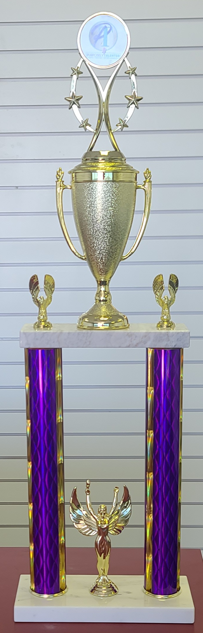 Tall two-column trophy with purple holographic pillars, gold cup topper, victory figure, and star riser on white marble base