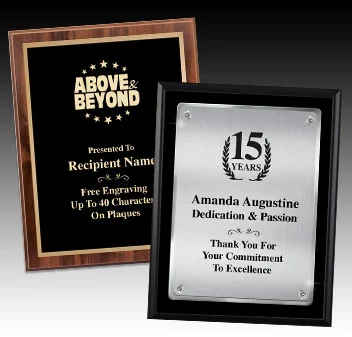 Two corporate award plaques are displayed side by side. The plaque on the left has a wood finish with a black and gold plate that reads "ABOVE & BEYOND" at the top, followed by space for a recipient name and a note about free engraving up to 40 characters. The plaque on the right has a sleek black frame with a silver plate, featuring a "15 YEARS" emblem at the top, followed by the name "Amanda Augustine" and text recognizing dedication and passion, with a thank-you message at the bottom.